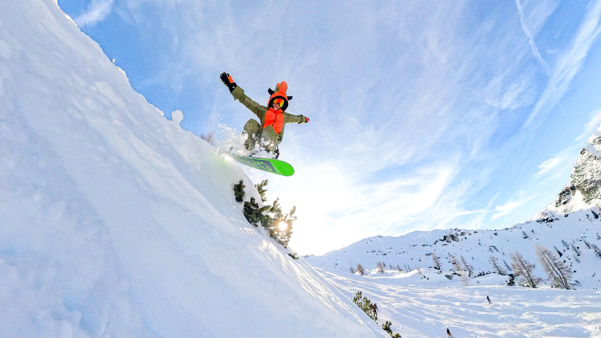 Child in an orange KHAMONDO snowsuit mid-air on a snowboard jump.