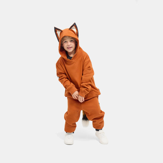 Child crouching in the rust-orange FOXDO Sweatsuit, hood up with fox ears, hoodie and joggers worn as a set.